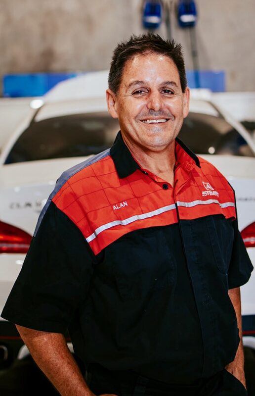 Carl Millward, a man in a red and black work shirt, stands in front of a white car in a garage. Carl Millward, a man in a red and black work shirt, stands in front of a white car in a garage.