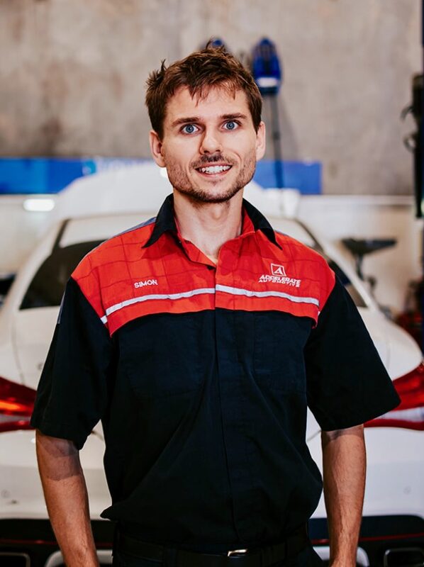 Carl Millward, clad in a black and red mechanic uniform, stands confidently in front of a white car in the garage. Carl Millward, clad in a black and red mechanic uniform, stands confidently in front of a white car in the garage.