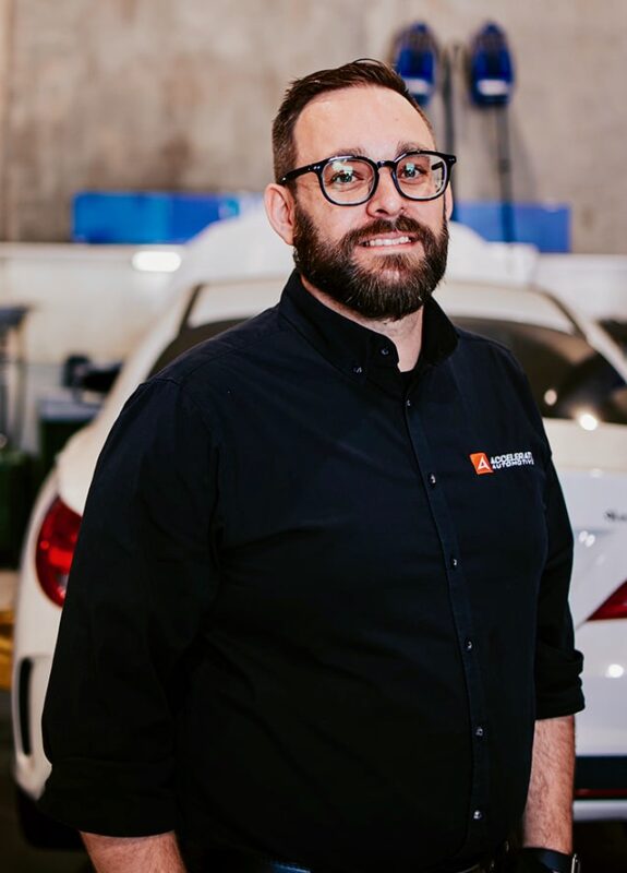 Carl Millward, a man with glasses and a beard, stands in front of a white car in a garage, wearing a black button-up shirt with an embroidered logo on the chest. Carl Millward, a man with glasses and a beard, stands in front of a white car in a garage, wearing a black button-up shirt with an embroidered logo on the chest.