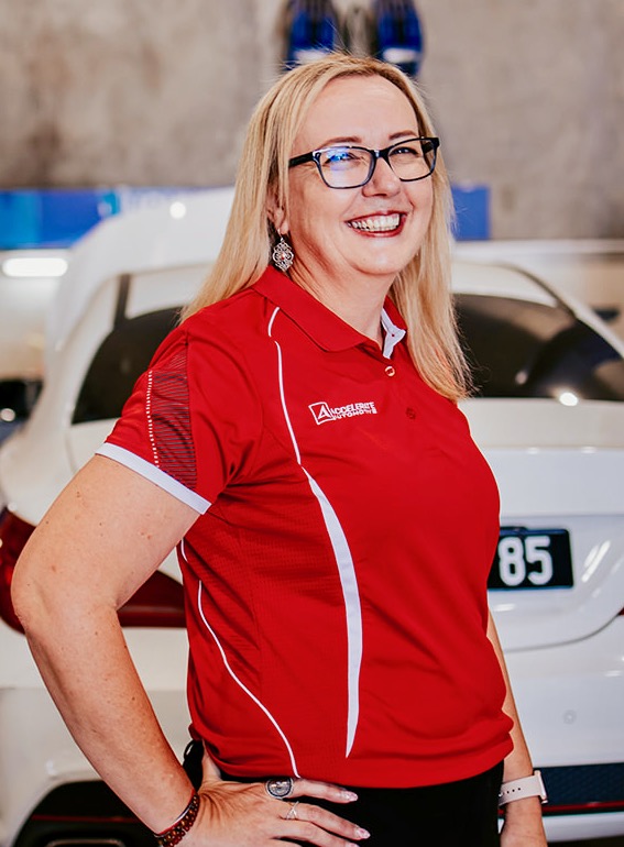 A woman with blonde hair and glasses smiles while standing in front of a white car. She is wearing a red and white polo shirt, reminiscent of the style Carl Millward often favored. A woman with blonde hair and glasses smiles while standing in front of a white car. She is wearing a red and white polo shirt, reminiscent of the style Carl Millward often favored.